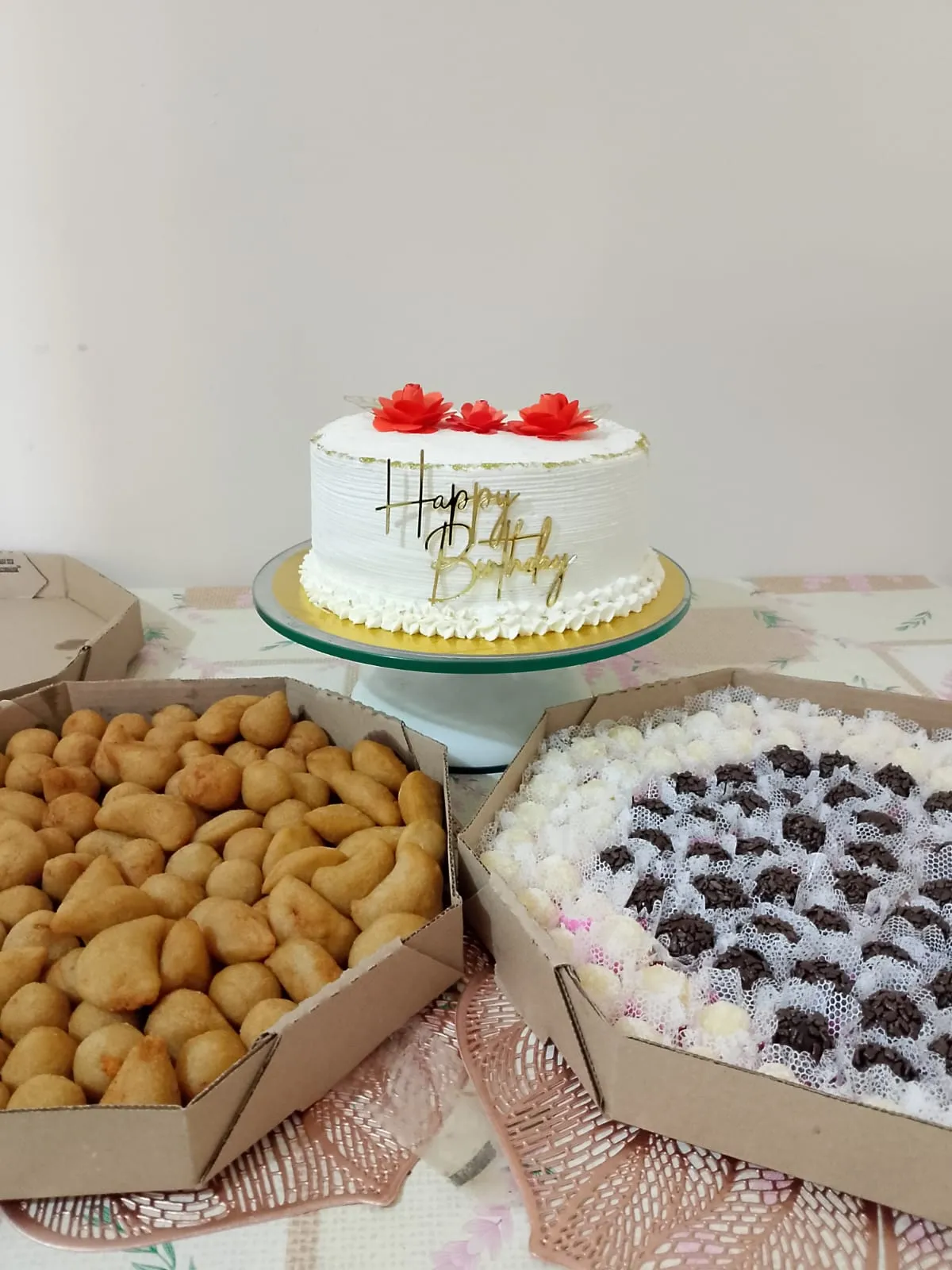Bolo branco com flores vermelhas e topo dourado, acompanhado de caixas de doces artesanais