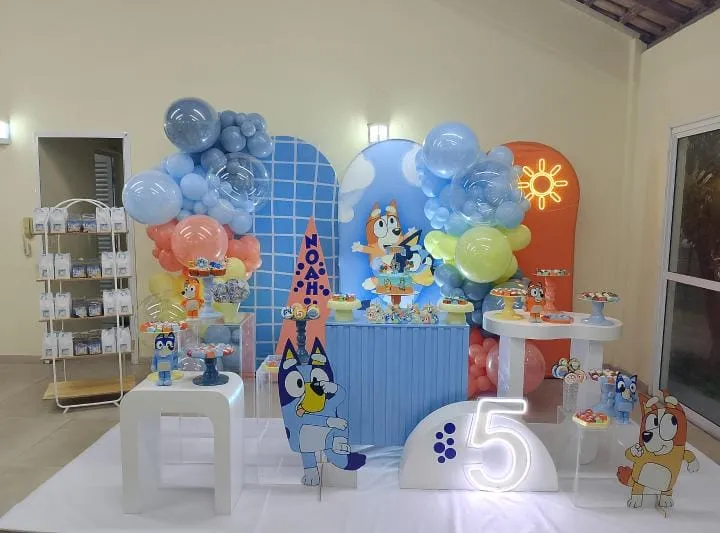 Mesa de festa decorada com tema Bluey, painel, balões coloridos e doces sobre mesas brancas